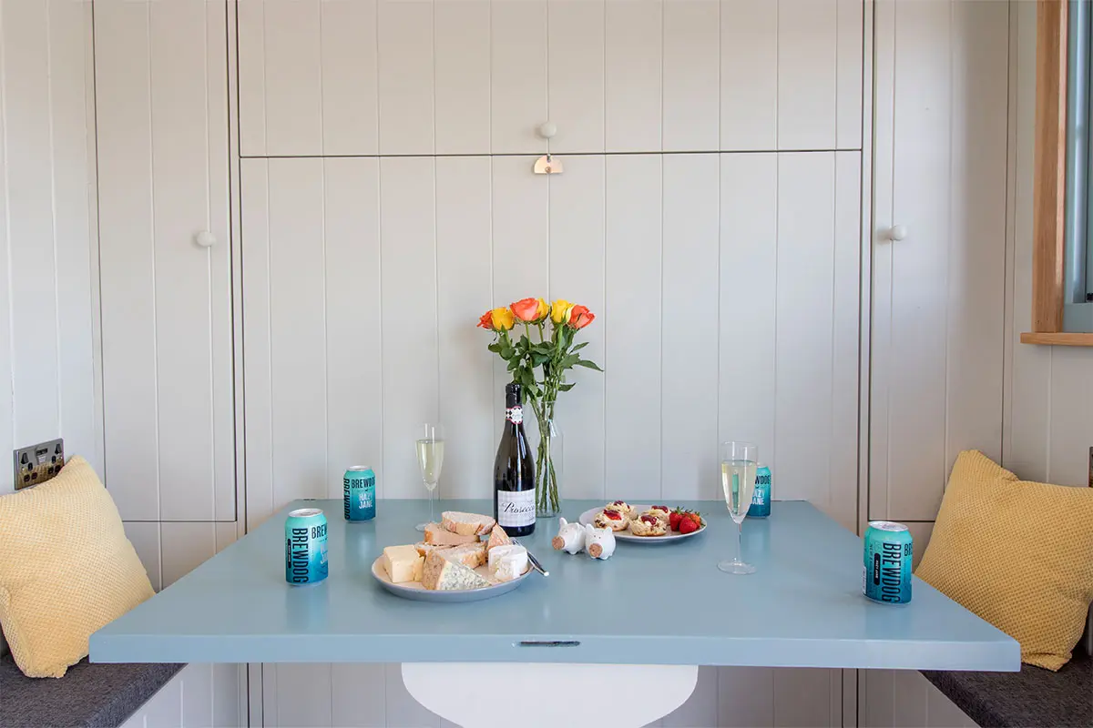 A blue table and flowers in a shepherds hut with a plate of cheeses and bread, another plate of scones and strawberries, 2 glasses of champagne and some cans of Brewdog